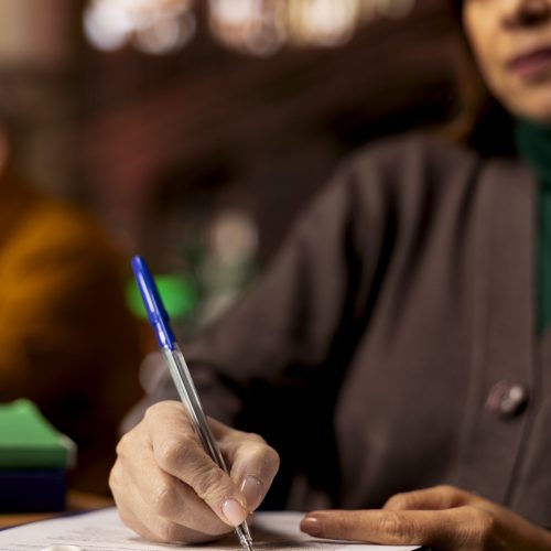 Adult learner transcribes information during a lecture in a university library, surrounded by academic textbooks and archived documents. Higher education personal goals on campus. Close up.